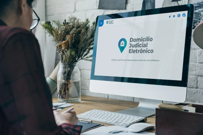 Pessoa sentada em frente a um computador acessando a plataforma “Domicílio Judicial Eletrônico”, com a interface do sistema exibida na tela. Sobre a mesa, há um teclado, um mouse, papéis e um vaso com galhos secos decorativos.