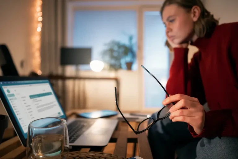 Pessoa trabalhando em home office à noite, segurando os óculos ao lado do notebook com a caixa de e-mail aberta; cena de foco, produtividade.