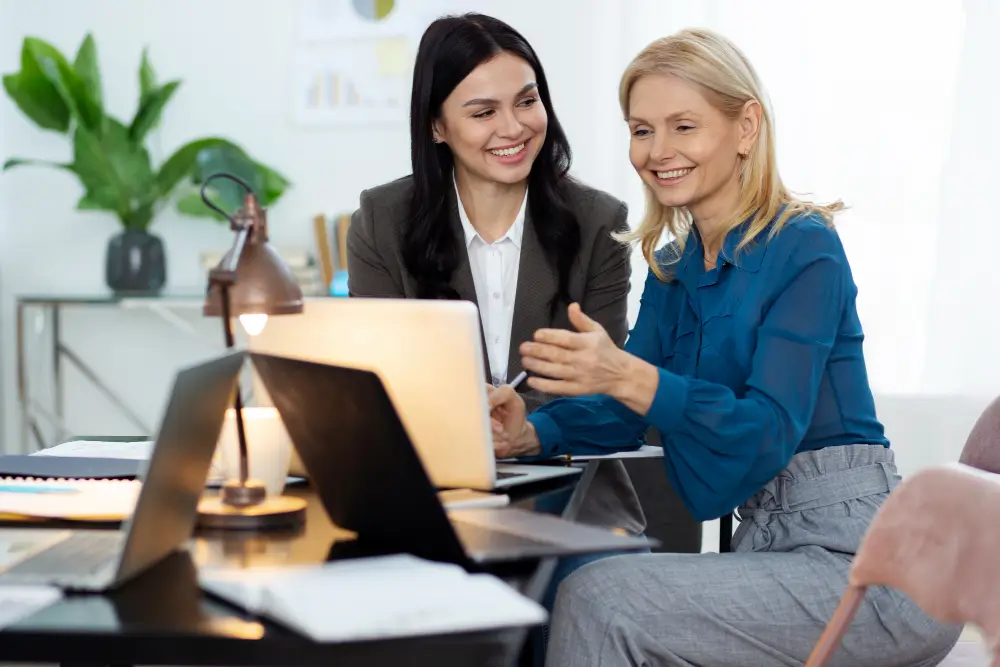 Duas mulheres em ambiente corporativo sorrindo e discutindo ideias diante de um laptop.